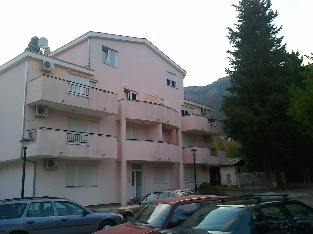 Three-story pink residential house with balconies in Baošići, Montenegro, against a backdrop of mountains and conifer trees.