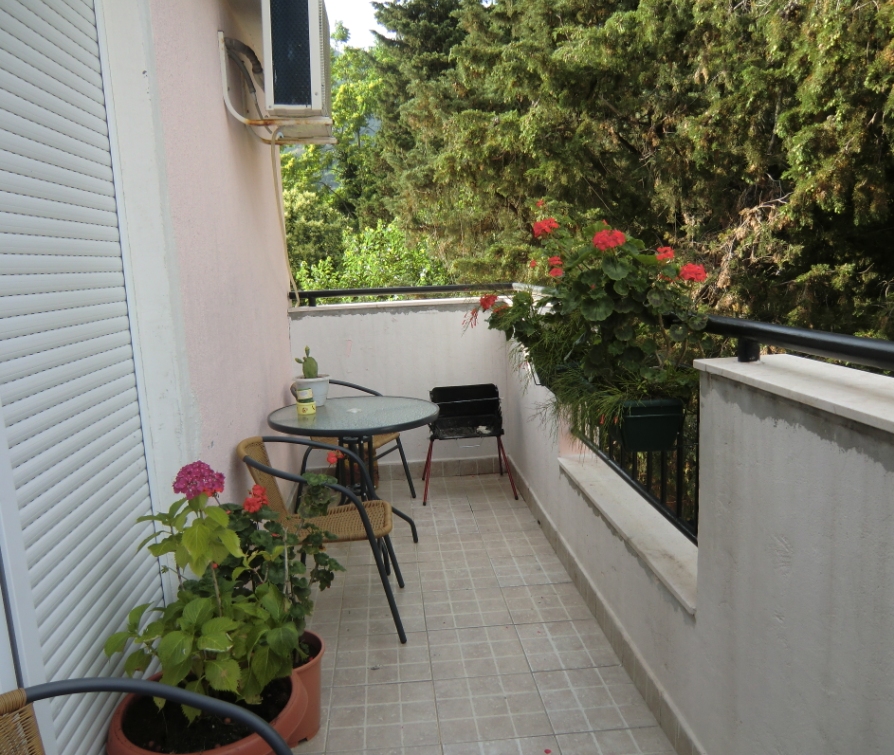 Cozy balcony with furniture, flowers, and a grill in an apartment in Baošići, Montenegro, overlooking a green garden.