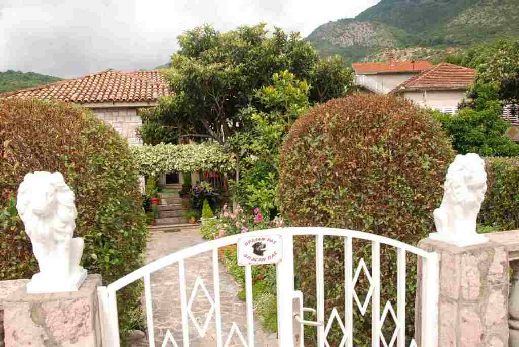 Eingang zum Haus in Kamenari, Montenegro: weiße Tore mit Löwenstatuen, gepflegter Garten und Bergblick.