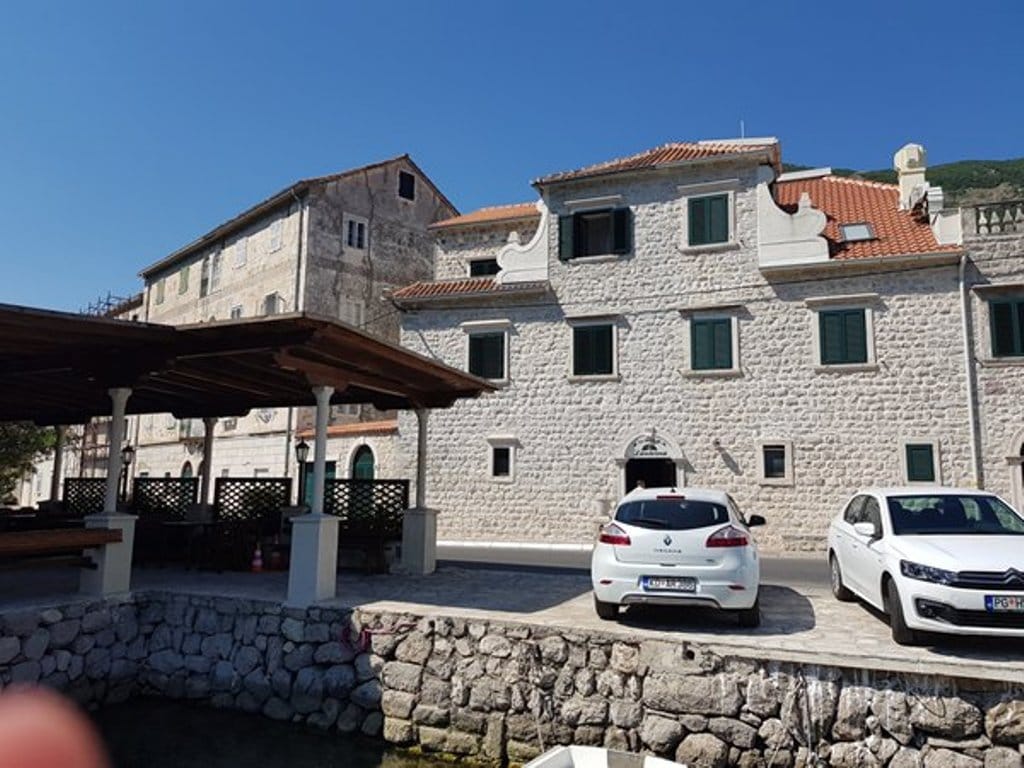 Karadağ, Prčanj’de deniz kenarında teraslı ve binanın cephesinin önünde park etmiş araçları olan geleneksel taş villa.