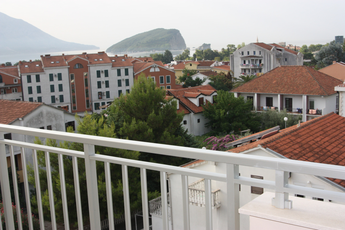Pogled sa balkona stana u Budvi, Crna Gora: panorama grada, crveni krovovi kuća, zelenilo i ostrvo Svetog Nikole.