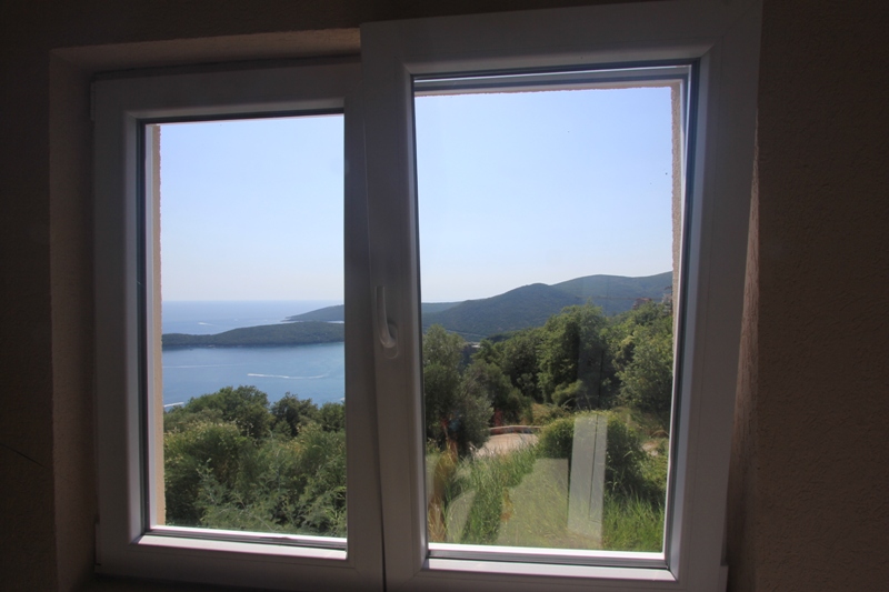 Blick aus einem Kunststofffenster auf das Meer und die Berge in einem Haus in Seoce, Montenegro: Panoramablick mit Grün und Küste.
