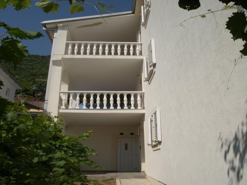 House facade in Baošići, Montenegro: light-colored walls, two open balconies with balustrades, and an entrance door.