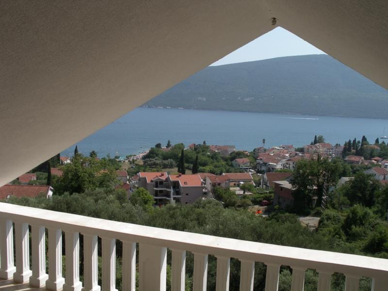 View from a house balcony in Baošići, Montenegro: a panorama of the sea, mountains, and coastal town through a triangular roof opening.