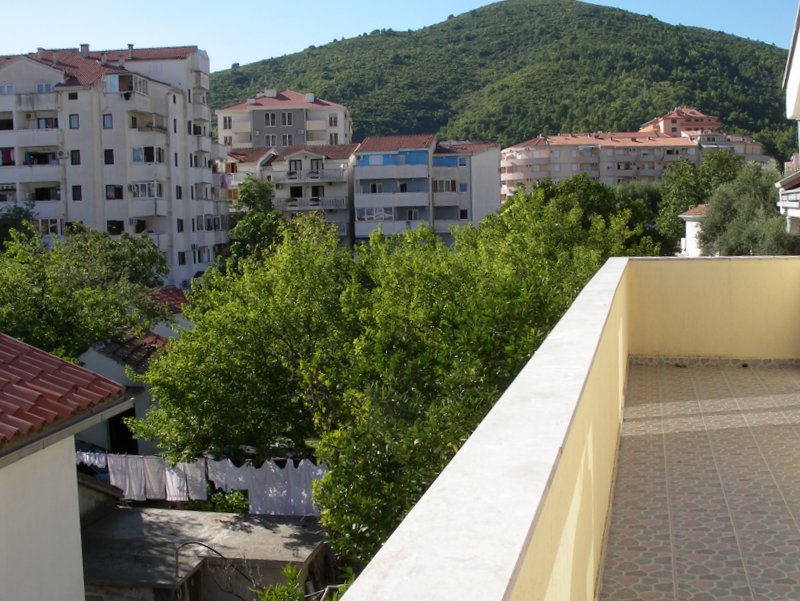 Pogled sa otvorenog balkona sa keramičkim pločicama na zelena stabla i stambene zgrade u Budvi, Crna Gora.