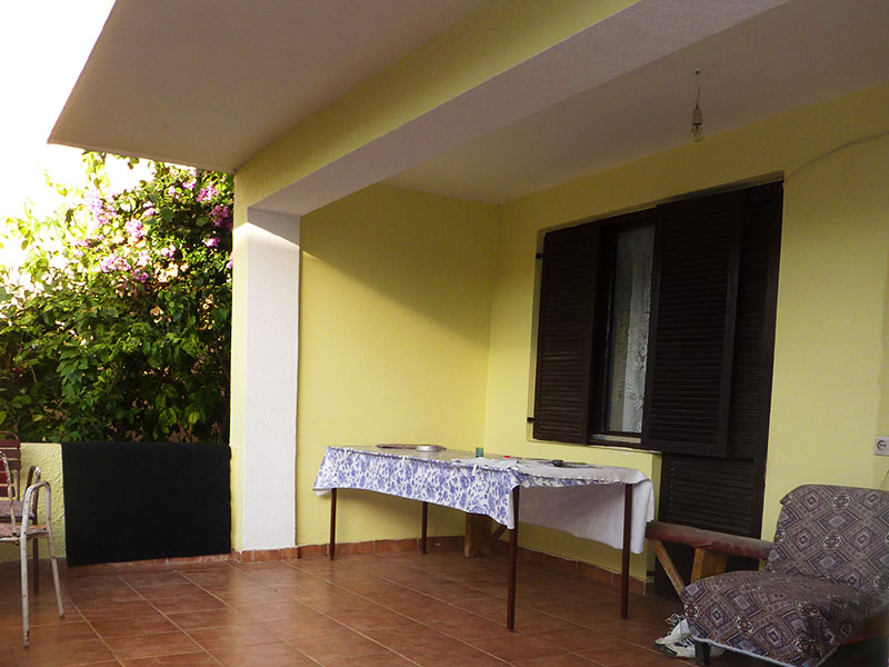 Überdachte Terrasse eines Hauses in Dobri Vody, Montenegro: Fliesenboden, Tisch mit weißer Tischdecke und Blick auf den blühenden Garten.