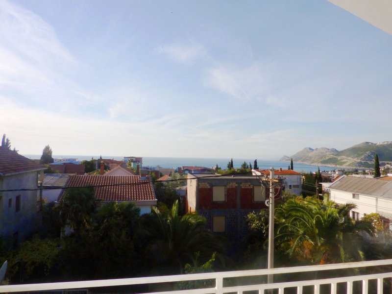 Blick auf das Meer und die Berge vom Balkon des Hauses in Dobri Vody, Montenegro: Panorama des Ferienortes mit Palmen und Ziegeldächern.