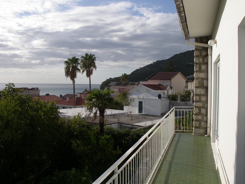 Blick vom Balkon eines Hauses in Petrovac, Montenegro: Panorama auf das Meer, Palmen, Berge und die Ziegeldächer der Stadt.