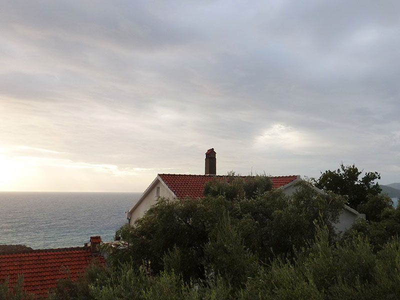 Meerblick und ein Haus mit rotem Ziegeldach in Sveti Stefan, Montenegro, umgeben von Grün an einem bewölkten Tag.