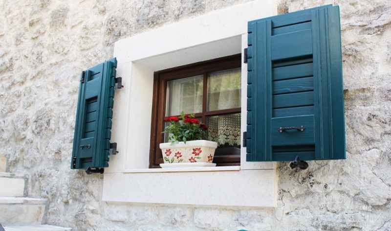Fenster eines Steinhauses in Strp, Montenegro, mit grünen Holzläden und einem Blumentopf auf der Fensterbank.