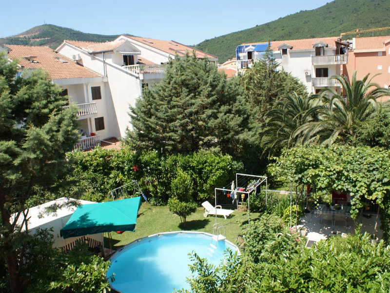 Budva, Karadağ’daki otelin havuz ve yemyeşil bahçe manzarası: şezlonglu dinlenme alanı, teras ve dağ manzarası.