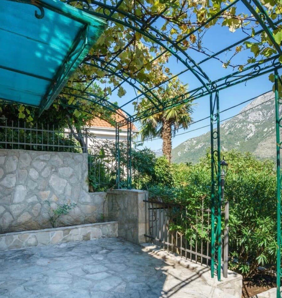 Terasa sa kovanim nadstrešnicom i vinovom lozom na vili u Stolivu, Crna Gora, s pogledom na planine i zeleni vrt.