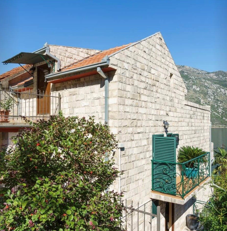 Kamena vila u Stolivu, Crna Gora: fasada od prirodnog kamena, zelene škure i balkon s pogledom na more i planine.