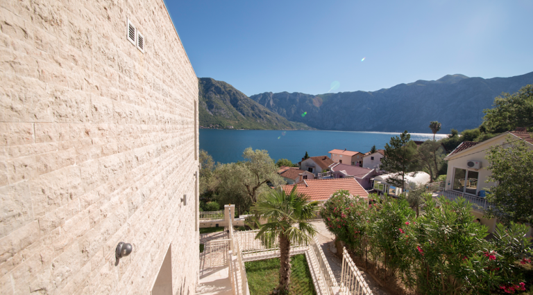 Blick auf die Bucht von Kotor und die Berge von der Terrasse eines Hauses in Stoliv, Montenegro: Steinfassade und gepflegter Garten mit Palme.