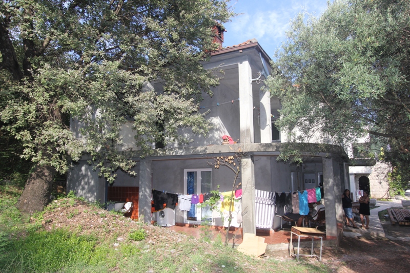 Zweistöckiges Haus mit Terrasse und Garten auf einem Grundstück in Budva, Montenegro: Außenansicht der Fassade zwischen Bäumen.