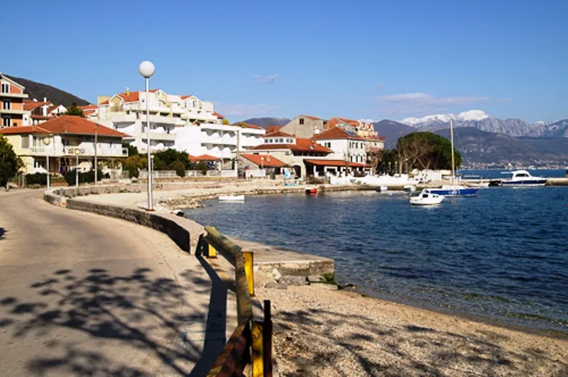 Blick auf ein Mini-Hotel in Đenovići, Montenegro: Küstenstraße, Strand, Yachten und schneebedeckte Berge im Hintergrund.