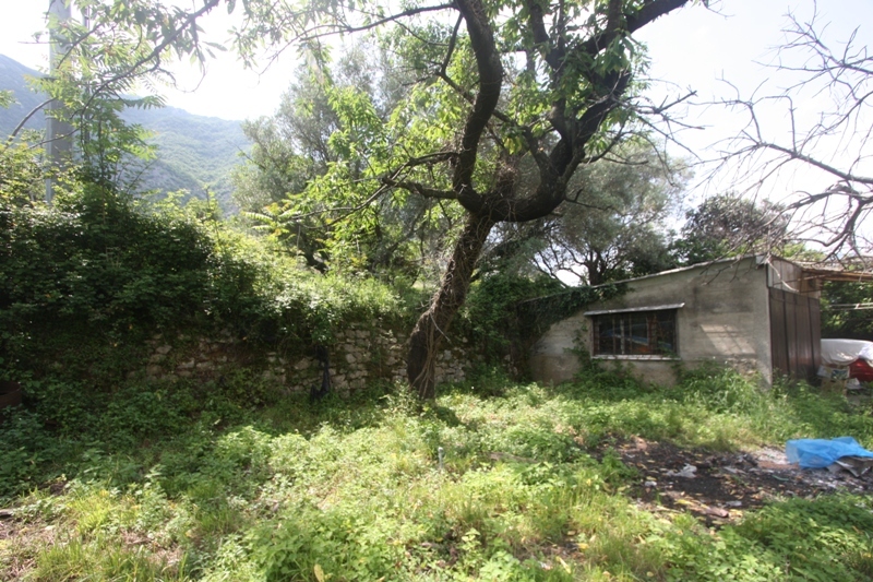 Land plot with a stone wall and an old house in Prčanj, Montenegro, surrounded by greenery and mountain scenery.