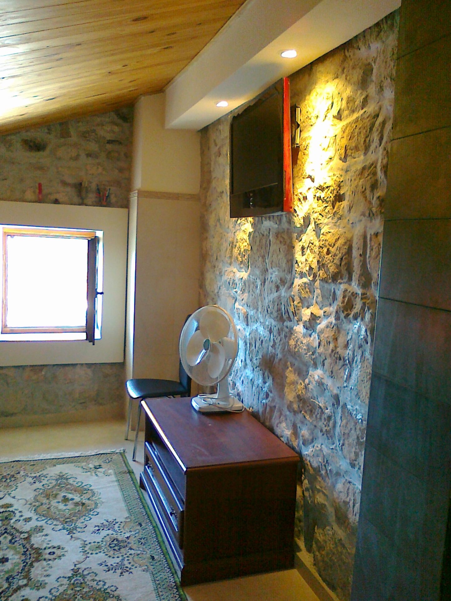 Wohnzimmer in einem Haus in Mišići, Bar, Montenegro: Steinwand, Holzdecke, TV-Möbel und Teppich im Interieur.