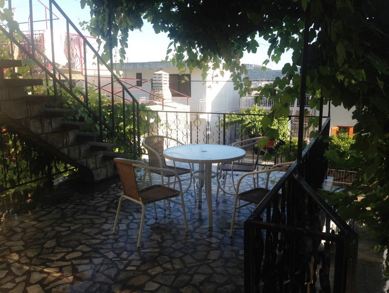 Ein schattiger Außenbalkon mit Tisch, Stühlen und Steinplattenboden, umgeben von Grün.