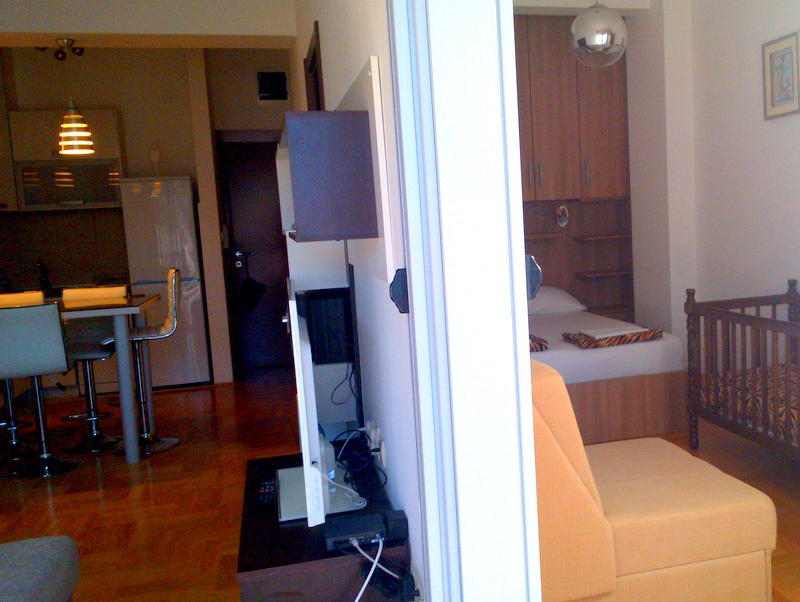 Blick aus dem Zimmer in Richtung Küchenbereich und einem Schlafzimmer mit Bett und Kinderbett.