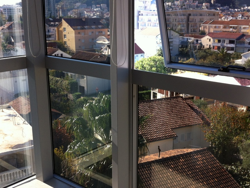 Blick auf Stadtgebäude und Bäume von einem Balkon mit großen Fenstern.