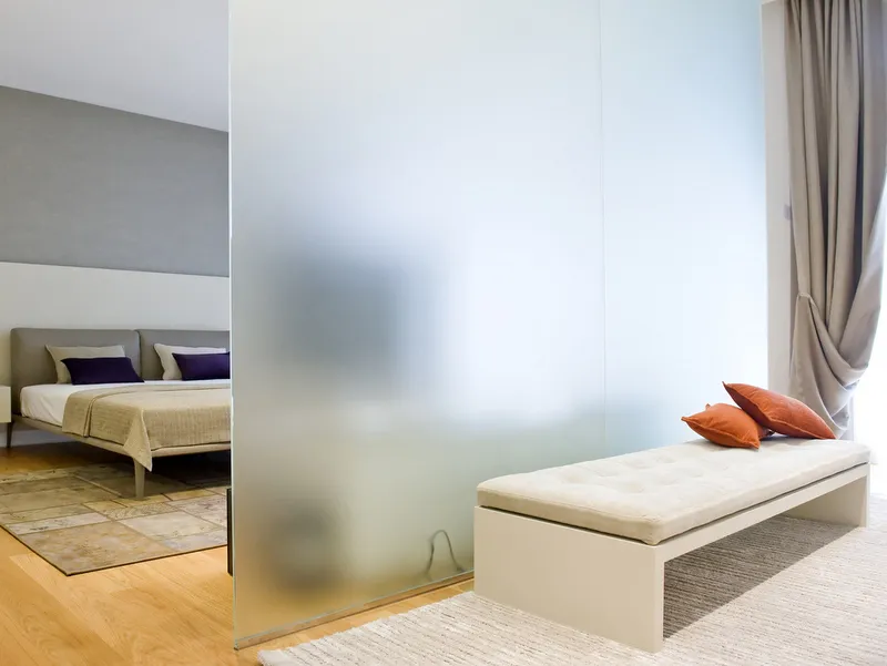 Ein modernes Schlafzimmer mit einer Milchglastrennwand und einer Bank mit orangen Kissen.