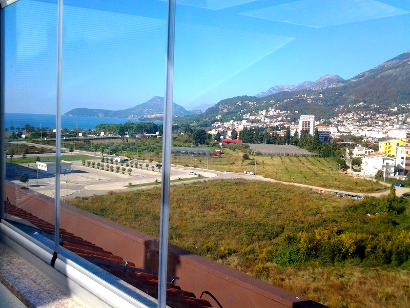Pogled sa zastakljenog balkona stana u gradu Bar, Crna Gora: panorama mora, planina i gradske infrastrukture.