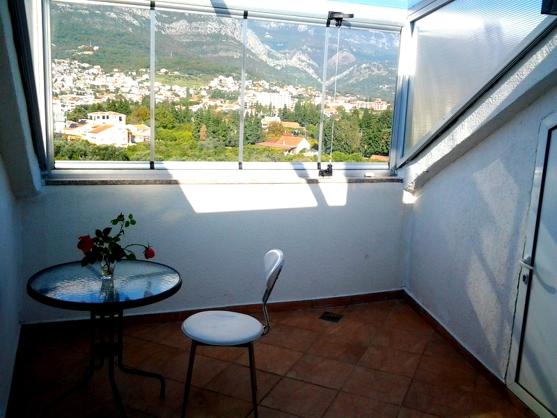 Balkon s panoramskim pogledom na planine i grad Bar, Crna Gora, sa staklenim stolom i stolicom u svetlom stanu.