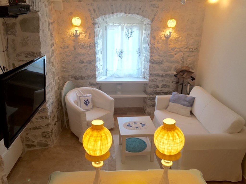 Udoban dnevni boravak sa kamenim zidovima u vili u Perastu, Crna Gora: beli nameštaj, meko svetlo i pogled kroz prozor.