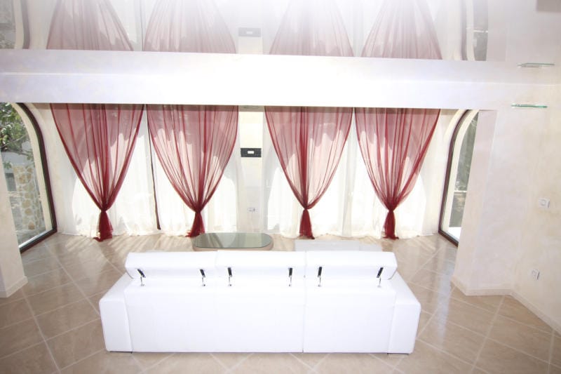 Salon de la villa à Lastva, Monténégro : canapé blanc, baies vitrées avec rideaux rouges et plafond brillant.