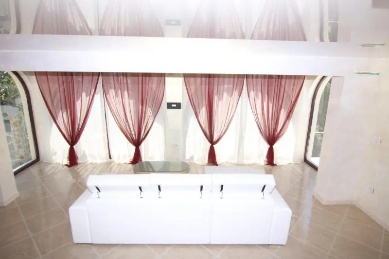 Salon de la villa à Lastva, Monténégro : canapé blanc, baies vitrées avec rideaux rouges et plafond brillant.