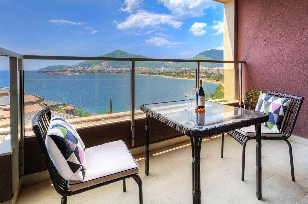 Balcon avec vue sur la mer et les montagnes dans un appartement, Bečići, Monténégro : chaises en osier, table en verre et baies vitrées panoramiques.