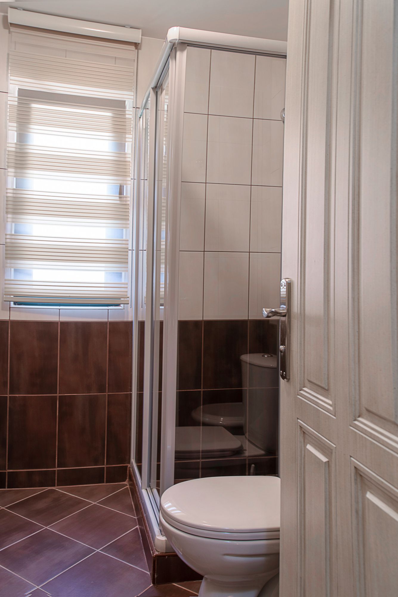 Salle de bain dans une maison de ville à Lastva, Monténégro : cabine de douche, carrelage clair et fenêtre avec stores.