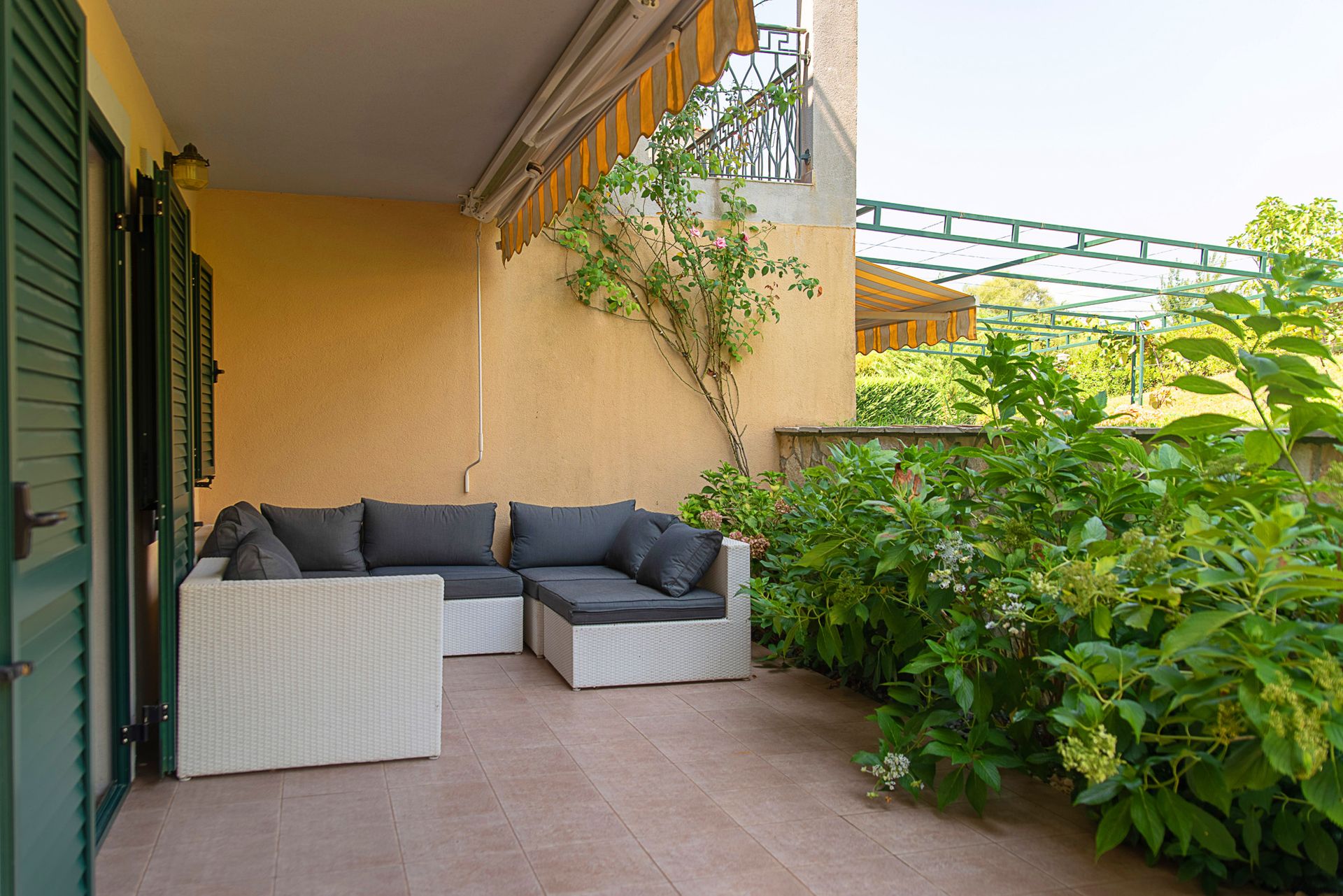 Terrasse confortable avec mobilier en osier et jardin dans une maison de ville, Lastva, Monténégro. Balcon ensoleillé avec auvent et verdure.