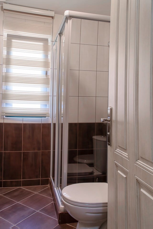 Salle de bains dans une maison de ville à Lastva, Monténégro : cabine de douche, carrelage clair et fenêtre avec stores.