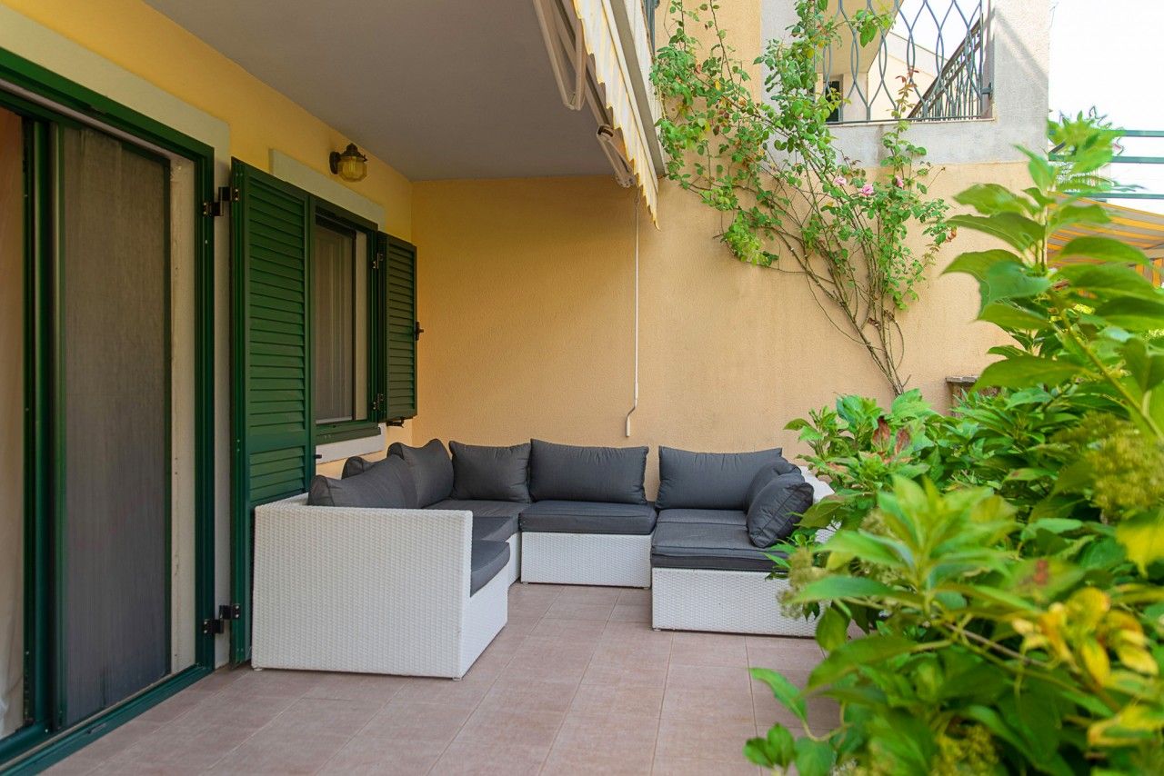 Terrasse cosy avec canapé en osier dans une maison de ville à Lastva, Monténégro : sol carrelé, volets verts et jardin.