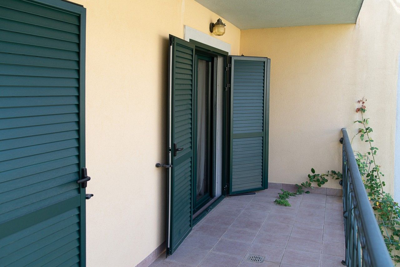 Balcon d’une maison de ville à Lastva, Monténégro : sol carrelé, volets verts sur les portes et garde-corps en fer forgé avec des plantes.
