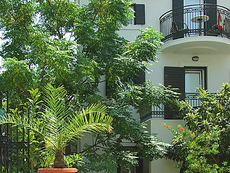Fassade des Hotels in Petrovac, Montenegro: Balkon mit schmiedeeisernem Geländer, grüner Garten mit Palme und Bäumen am Gebäude.