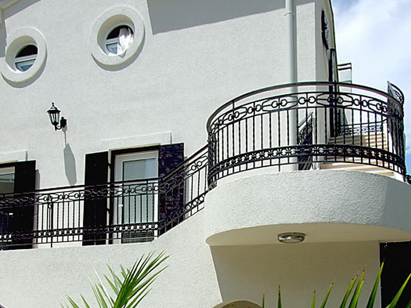 Fassade des Hotels in Petrovac, Montenegro: weiße Wände, runde Fenster und schmiedeeiserne Geländer auf der Terrasse des Gebäudes.