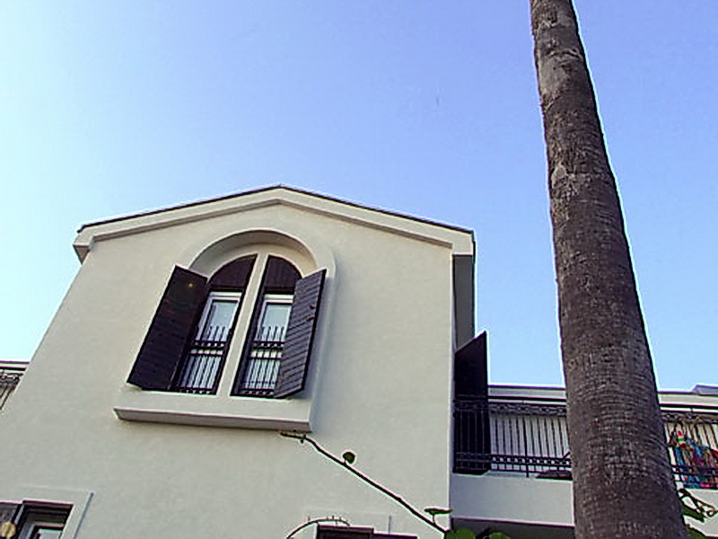 Fassade eines Hotels in Petrovac, Montenegro: weiße Wände, ein Bogenfenster mit dunklen Holzläden und eine hohe Palme.