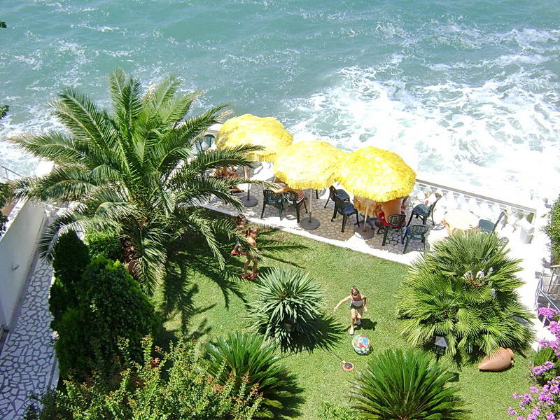 Blick von oben auf den grünen Hotelgarten in Sutomore, Montenegro, mit Palmen, gelben Sonnenschirmen am Meer und einer Ruhezone auf dem Rasen.