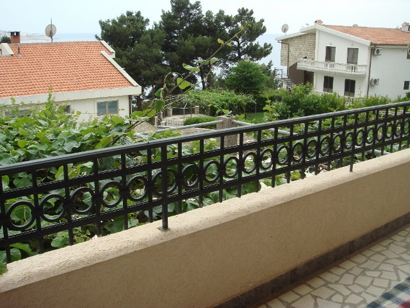 Ein Balkon mit Metallgeländer und beiger Wand, mit Blick auf Häuser, Bäume und das Meer.