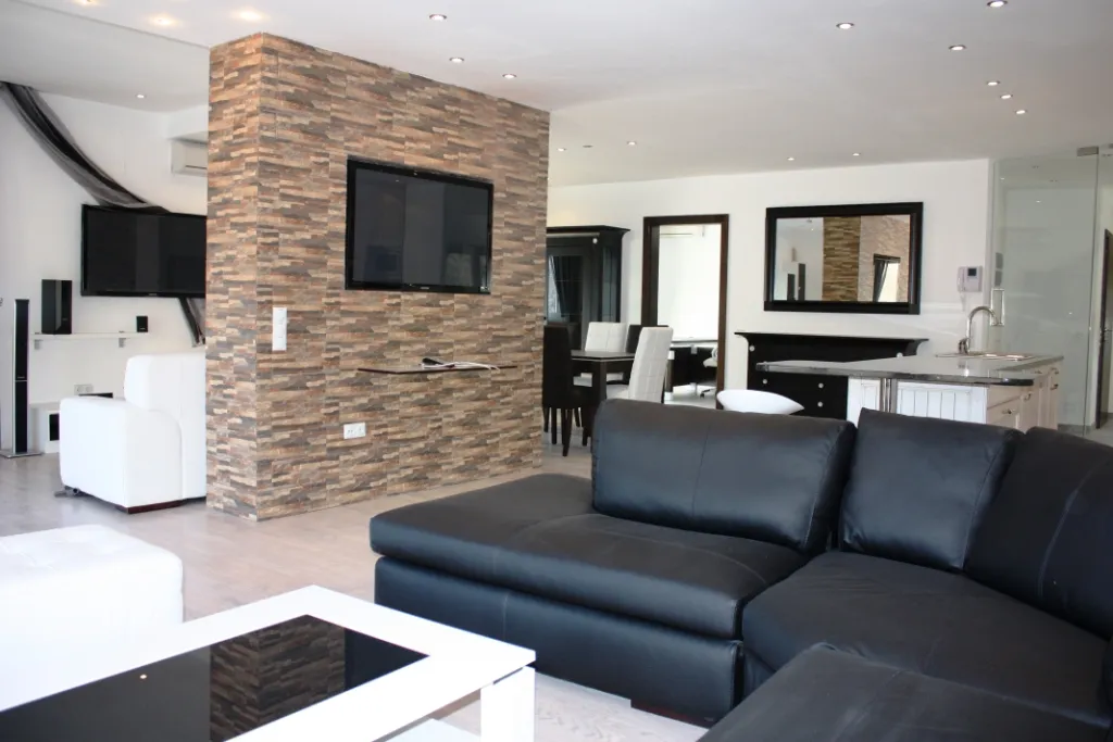 Geräumiges Wohnzimmer mit Ziegelsäule, schwarzem Ledersofa und Küchenbereich im Hintergrund.