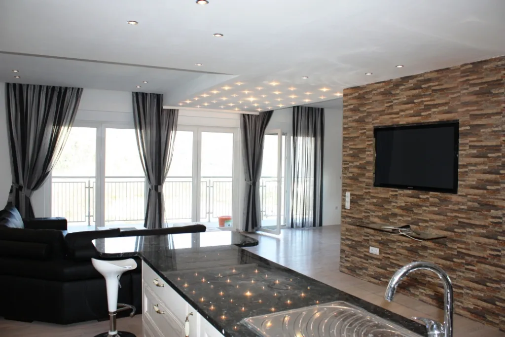 Modernes Wohnzimmer mit Steinakzentwand, Fernseher, großen Fenstern und einer Kücheninsel.