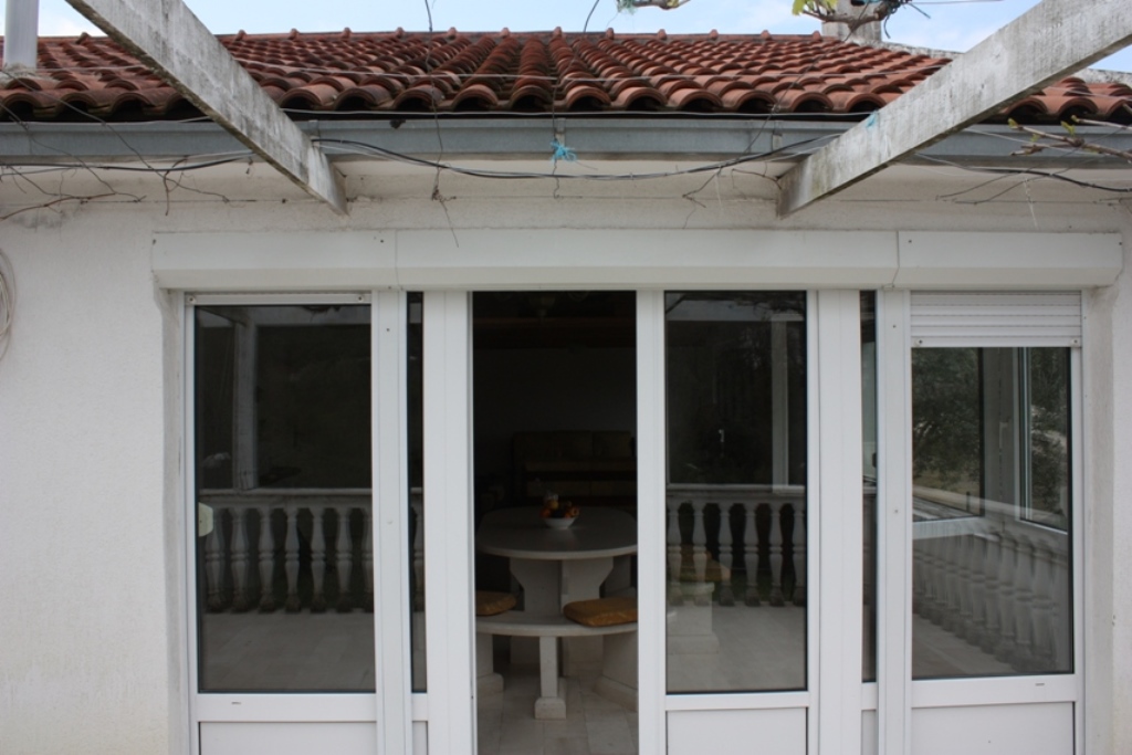 Fassade des Hauses in Bigovo, Montenegro: Terrasse mit Steintisch, Panoramafenster und Ziegeldach mit Pergola.