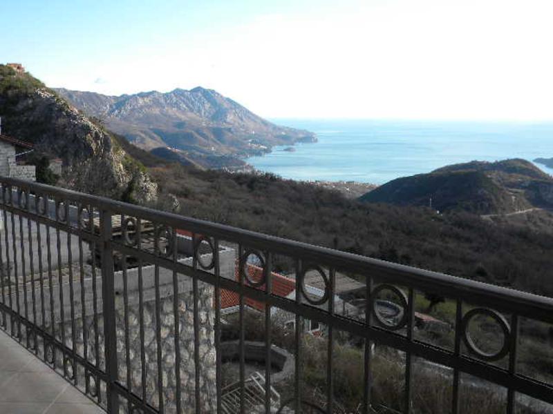 Panoramablick auf das Meer und die Berge vom Balkon eines Hauses in Budva, Montenegro, mit schmiedeeisernem Geländer und Steinverkleidung der Fassade.