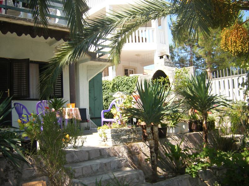 Gemütliche Terrasse eines Hauses in Tivat, Montenegro, mit Gartenmöbeln, Palmen und steinverkleideter Fassade im Grünen.