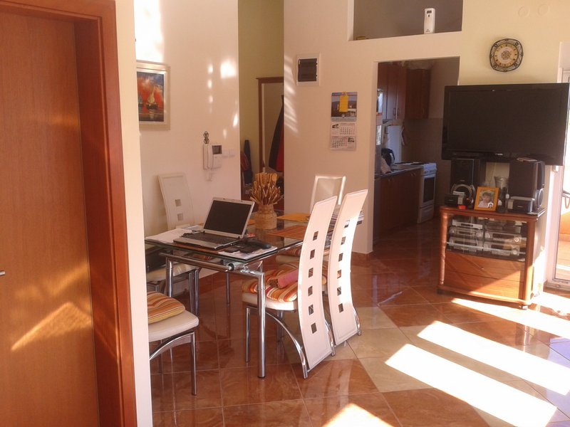 Wohnzimmer mit Essbereich in einer Wohnung in Igalo, Montenegro: Glastisch, weiße Stühle und Fliesenboden.