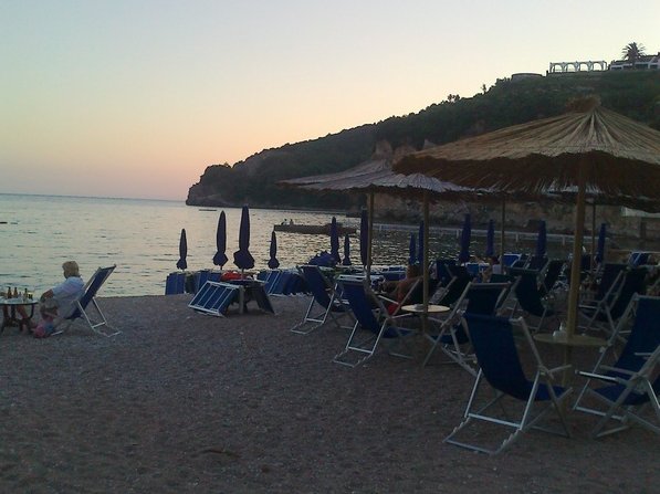 Blick auf den Strand mit Liegestühlen und Strohhüten am Meer in Budva, Montenegro, vor der malerischen felsigen Küste.