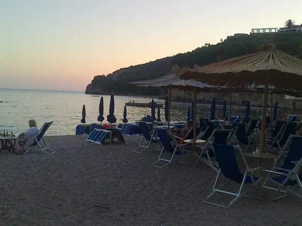 Blick auf den Strand mit Liegestühlen und Strohhüten am Meer in Budva, Montenegro, vor der malerischen felsigen Küste.